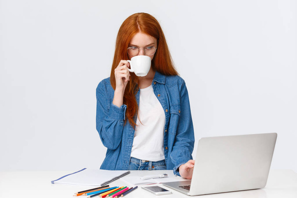 Серьезный вид рыжеволосая женщина дизайнер, офисный работник нет времени на кофе-брейк, потягивая из чашки, глядя на ноутбук, как работает над важным проектом, стоять белый фон
 - Фото, изображение