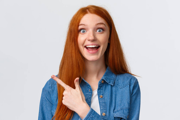 Close-up portrait excited happy ginger girl, foxy hair and blue eyes, smiling upbeat with amazement and joy, pointing finger left, attend amusing cool party, telling about it, white background - Photo, Image