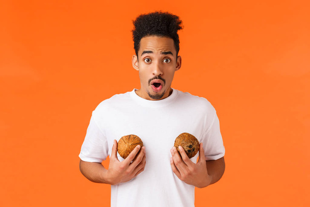Worried and concerned african-american male tourist in white t-shirt, holding coconuts on breast, gasping anxious and nervous, looking camera shocked, standing orange background - Photo, image