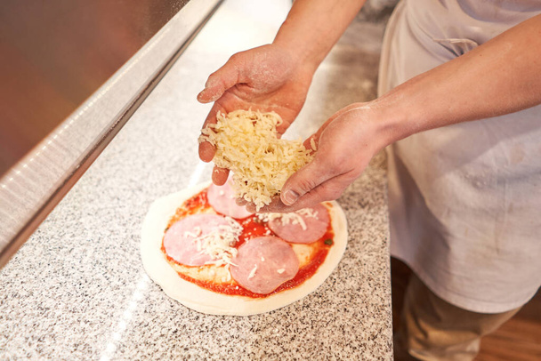 Приготування піци. готує сирні інгредієнти на тісті. крупним планом рука шеф-кухаря в уніформі білого фартуха готують на кухні
 - Фото, зображення