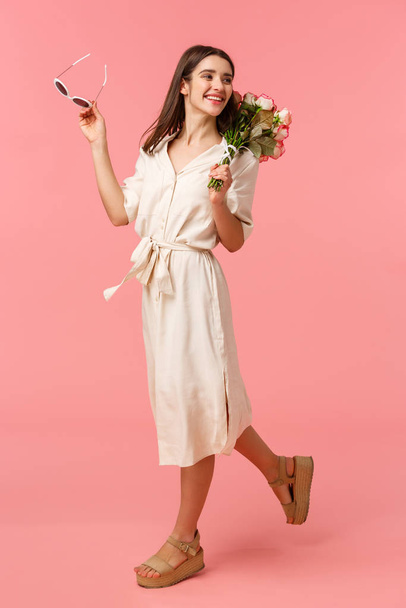 full-length vertical retrato despreocupado atraente feminino morena no vestido, andando ao longo da rua, recebendo belas flores, olhar para trás e sorrindo, passarela sobre fundo rosa
 - Foto, Imagem