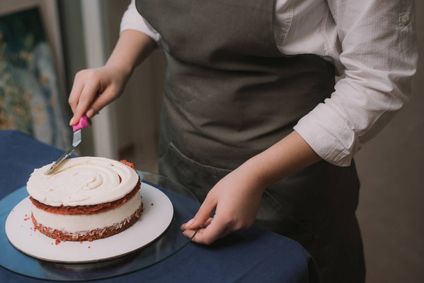 The confectioner levels the cream on the cake. The cook creates a cake, collects it and covers it with cream and then levels it with a spatula and a scraper - Foto, afbeelding