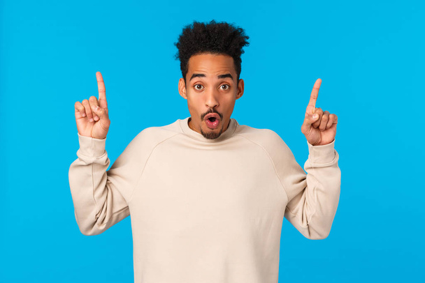 Have you seen it. Attractive impressed african-american guy with moustache, afro haircut, saying wow, telling about event, product advertising, pointing fingers up over blue background - Zdjęcie, obraz