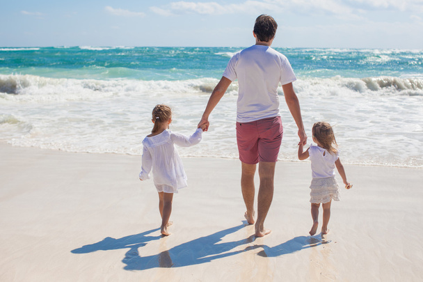 Вид на молодого отца и его восхитительных маленьких дочерей, гуляющих по белому песчаному пляжу в солнечный день
 - Фото, изображение