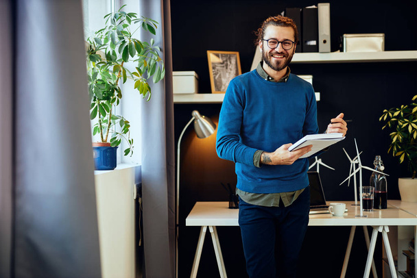 Молодой улыбающийся кавказский инновационный графический дизайнер стоит в своем кабинете и пишет идеи в блокноте
. - Фото, изображение