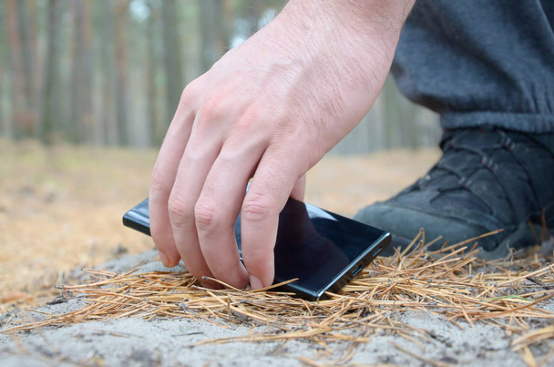 La mano masculina recogiendo el teléfono móvil perdido del suelo en el camino otoñal de abeto de madera. El concepto de encontrar una cosa valiosa y buena suerte
 - Foto, Imagen