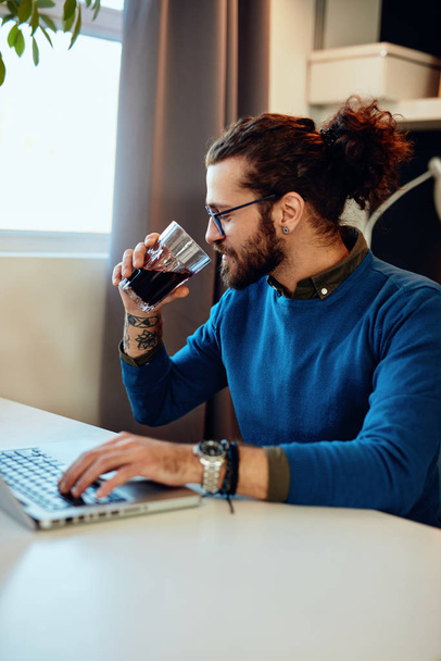 Attractive bearded caucasian entrepreneur sitting in his office, typing report on laptop and drinking fresh juice.ng in his office, - Foto, afbeelding
