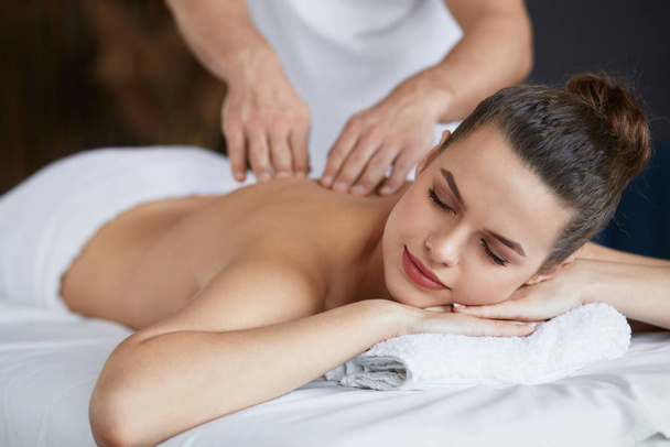 Jonge mooie vrouw genieten van rug en shouders massage in spa.Professionele massage therapeut is de behandeling van een vrouwelijke patiënt in appartement.Ontspanning, schoonheid, lichaam en gezicht behandeling concept.Home massage - Foto, afbeelding