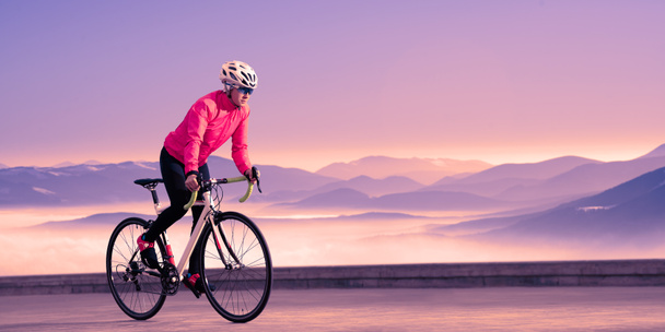 Νεαρή γυναίκα ποδηλάτης Riding Road Bike στα όμορφα βουνά στο μωβ ηλιοβασίλεμα. Περιπέτεια, Υγιεινός τρόπος ζωής, Αθλητισμός - Φωτογραφία, εικόνα