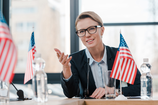 smiling diplomat in eyeglasses gesturing while talking near microphone and american flag  - Photo, Image