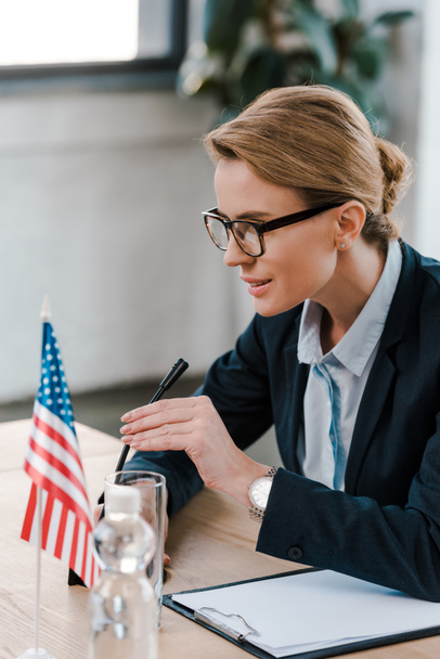 houkuttelevan diplomaatin valikoiva keskittyminen silmälaseihin, jotka koskettavat mikrofonia leikepöydän ja Amerikan lipun lähellä
  - Valokuva, kuva