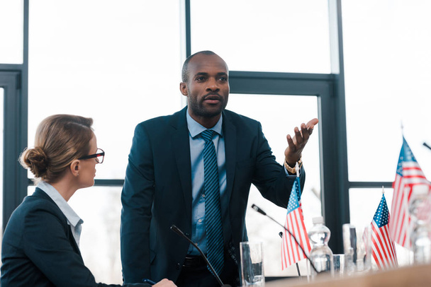 selectieve focus van aantrekkelijke diplomaat op zoek naar Afrikaanse Amerikaanse man gebaren tijdens het praten in de buurt vlaggen van Amerika  - Foto, afbeelding