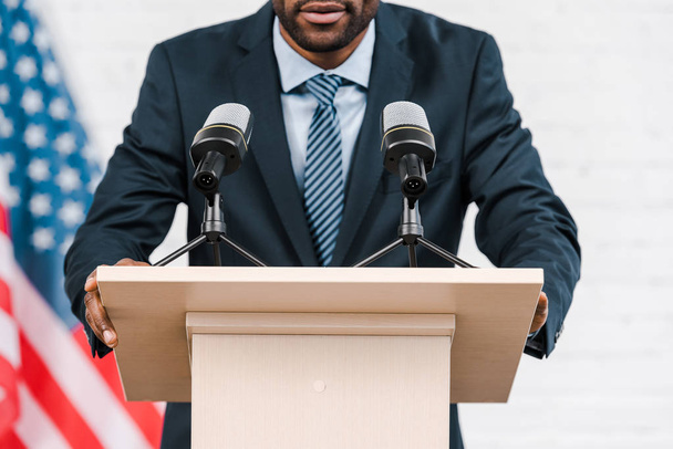 abgeschnittene Ansicht eines afrikanisch-amerikanischen Sprechers, der in der Nähe von Mikrofonen und amerikanischer Flagge spricht  - Foto, Bild