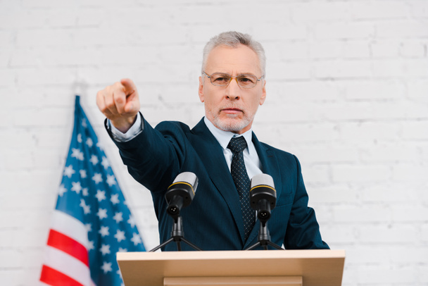 handsome speaker in glasses pointing with finger near microphones and american flag  - Fotoğraf, Görsel