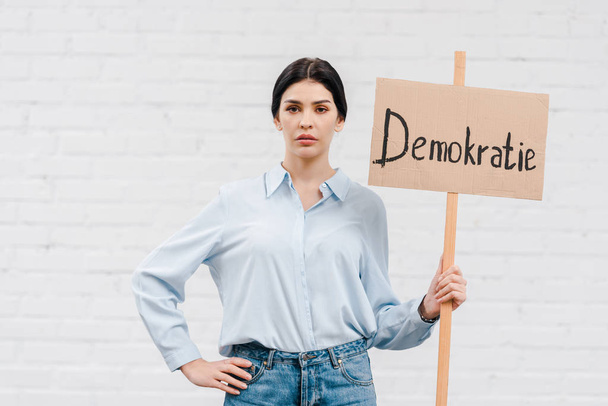 femme tenant une pancarte avec lettrage demokratie et debout avec la main sur la hanche près du mur de briques
  - Photo, image