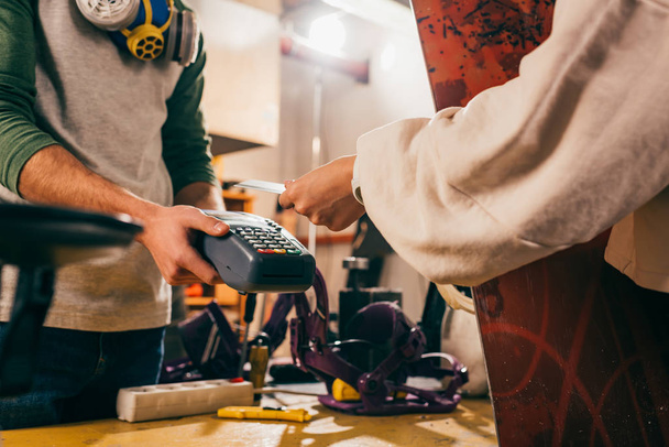 cropped view of worker holding terminal and woman paying with credit card and holding snowboard in repair shop  - Photo, Image
