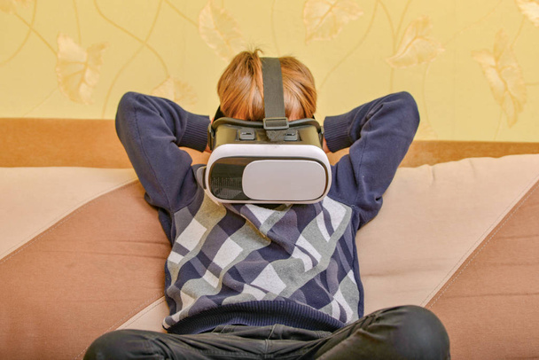 Chlapec v helmě virtuální reality sleduje video doma na gauči - Fotografie, Obrázek