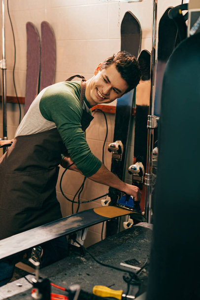 smiling worker waxing ski with wax iron in repair shop - Foto, Imagem