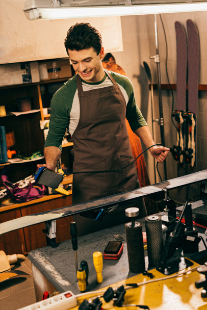 smiling worker waxing ski with wax iron in repair shop - Photo, Image