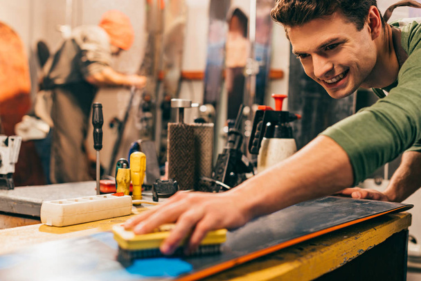 smiling worker using brush on snowboard in repair shop  - Фото, зображення