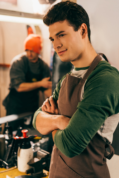 selective focus of smiling worker with crossed arms and his colleague on background in repair shop  - Фото, зображення
