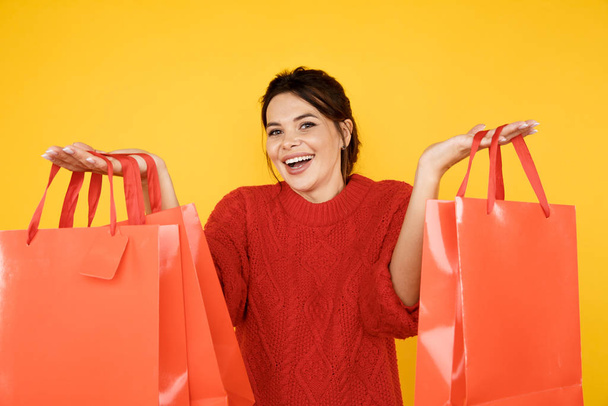 Szczęśliwa młoda dama w czerwonym swetrze z torbami na zakupy w żółtym studio. - Zdjęcie, obraz