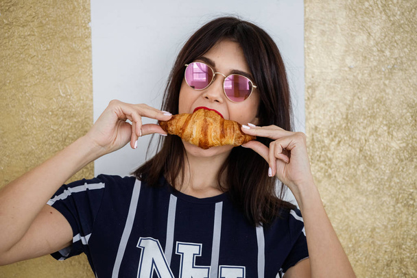 Beautiful brunette girl in glasses eating croissant at apartment interior  - Фото, изображение