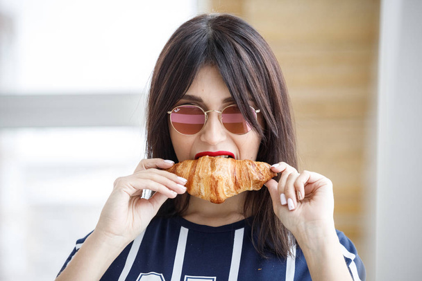 Beautiful brunette girl in glasses eating croissant at apartment interior  - Zdjęcie, obraz