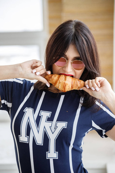 Beautiful brunette girl in glasses eating croissant at apartment interior  - Фото, изображение