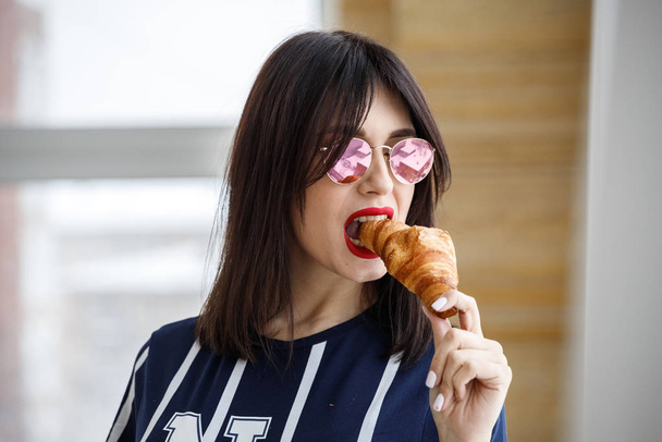 Beautiful brunette girl in glasses eating croissant at apartment interior  - Foto, Imagem