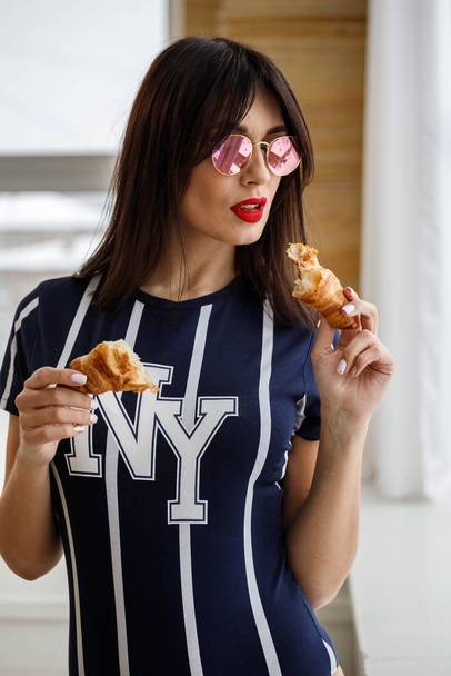 Beautiful brunette girl in glasses eating croissant at apartment interior  - Photo, image