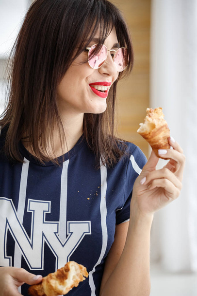 Beautiful brunette girl in glasses eating croissant at apartment interior  - Zdjęcie, obraz