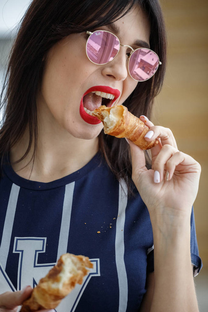 Beautiful brunette girl in glasses eating croissant at apartment interior  - Foto, imagen