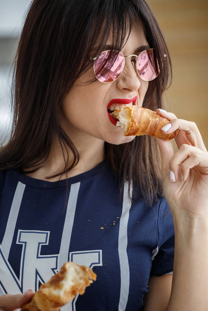 Beautiful brunette girl in glasses eating croissant at apartment interior  - Foto, afbeelding