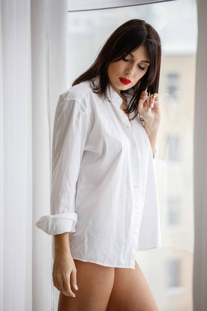 Attractive brunette in white shirt posing morning near window at morning time  - 写真・画像