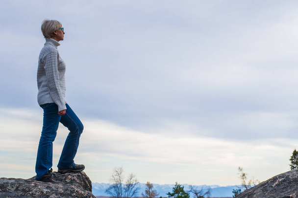 A brooding woman with glasses, dressed in a white sweater and jeans. Stands on top of a mountain against the sky and clouds. Travel concept and background with space for text. Dreams and loneliness. - Foto, imagen