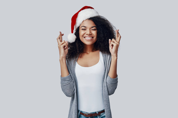 speelse jonge Afrikaanse vrouw in Santa hoed houden vingers gekruist en glimlachen terwijl staan tegen grijze achtergrond - Foto, afbeelding