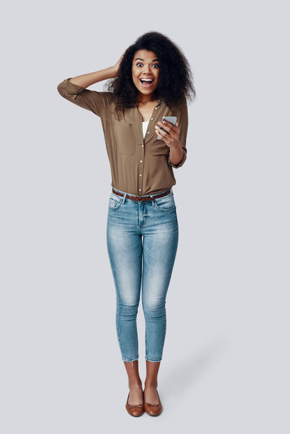 Longitud completa de la joven mujer africana sorprendida usando el teléfono inteligente y sonriendo mientras está de pie contra el fondo gris
 - Foto, Imagen