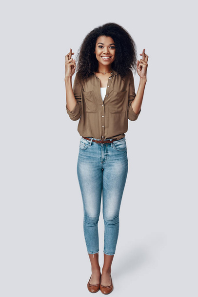 Longitud completa de joven mujer africana juguetona manteniendo los dedos cruzados y haciendo una cara mientras está de pie sobre fondo gris
 - Foto, Imagen