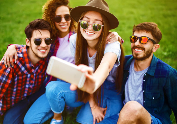 Group of smiling friends with phone sitting on grass and making selfie in summer park. Teenagers having fun together. Rest, fun, summer, technology and people concept. - Photo, image