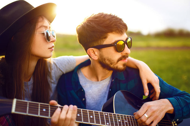 男は公園の芝生の上に座ってピクニックで自分の女の子にギターを演奏。夏の公園でパーティーを楽しむ若い人たち。休息、楽しみ、夏の時間を一緒にコンセプト. - 写真・画像