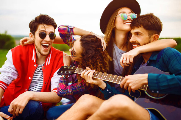 Ryhmä onnellisia ystäviä kitara hauskaa viettää vapaa-aikaa yhdessä puistossa istuu nurmella. Hän soittaa kitaraa. Kesäpuistossa juhlitaan. Lepo, hauska, kesäkonsepti
. - Valokuva, kuva
