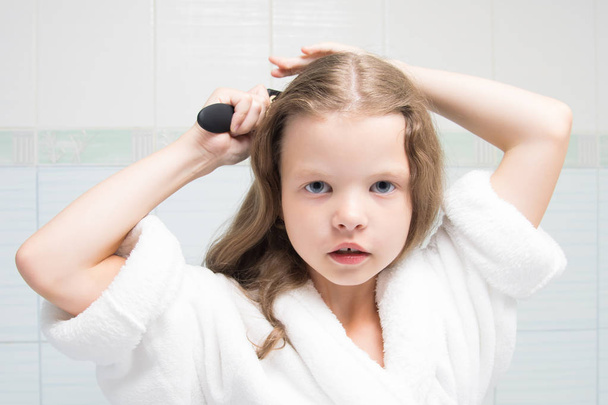 menina em um manto branco, no banheiro, penteando seu cabelo
 - Foto, Imagem