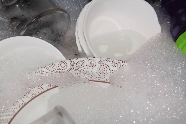 Geschirr in der Küche waschen. Schmutziges Geschirr, Teller und Gläser liegen in der Spüle aus weißem Schaum. auf dem Foto, weiße und graue Farben. - Foto, Bild