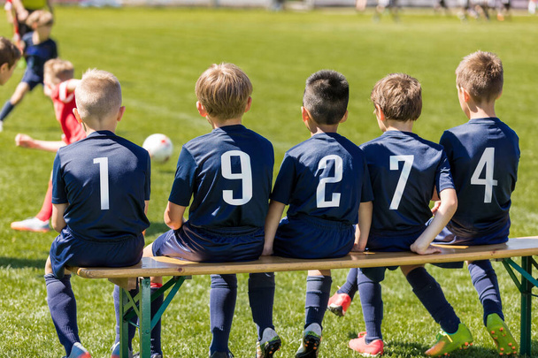 Amigos en un equipo de fútbol sentados en un banco de madera. Grupo de niños interraciales en un equipo deportivo escolar
 - Foto, Imagen