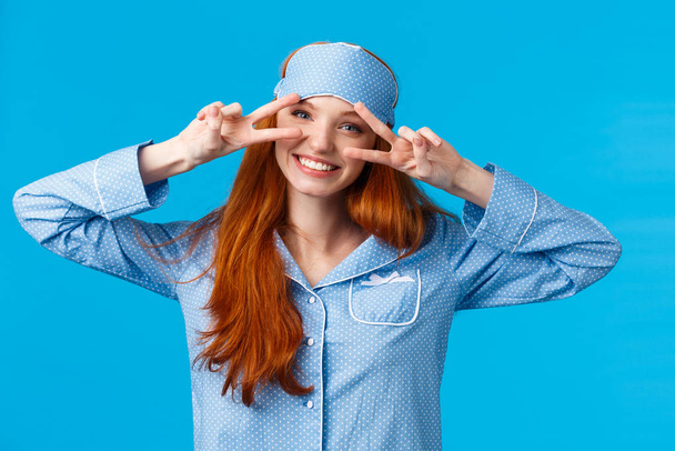 Energized cheerful girl slept well, wake up morning enthusiastic and excited, showing peace signs feeling optimistic smiling toothy, wearing sleep mask and nightwear, standing blue background - 写真・画像