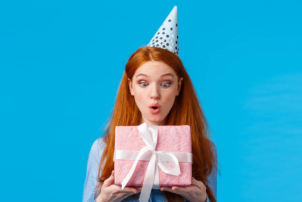 Wow amazing. Impressed and excited touched cute glamour redhead girl with freckles, folding lips thrilled, look at received pink wrapped b-day gift thankful standing happy blue background - Photo, Image