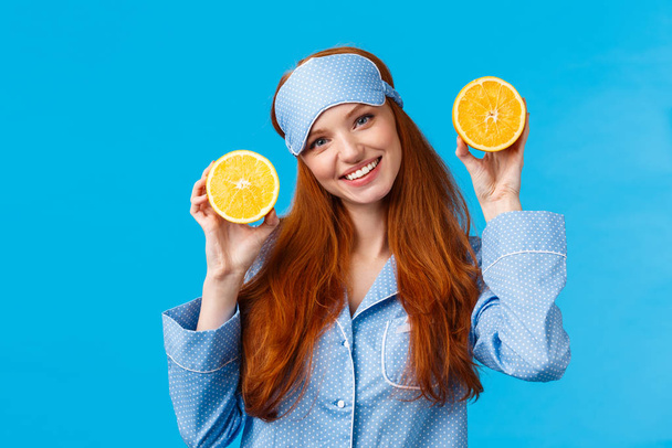 Dívka navrhuje ovoce k snídani. Roztomilá a něžná zrzka žena ve spánkové masce, pyžama, naklánět hlavu a držet dva plátky pomeranče, usmívat se, připravit šťávu, jíst zdravé ráno, modré pozadí - Fotografie, Obrázek
