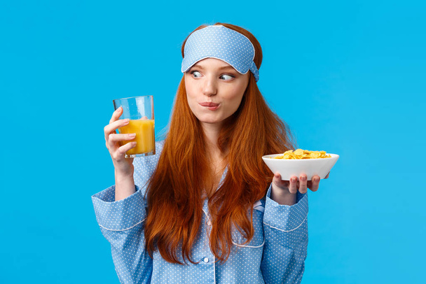 Beztrosko wyglądająca biała ślicznotka, studentka uśmiechnięta rozbawiona gapieniem się na sok pomarańczowy, trzymaniem płatków śniadaniowych, jedzeniem porannego posiłku, śniadaniem, stojącym niebieskim tle - Zdjęcie, obraz