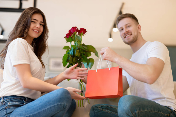 lovely couple sitting on the sofa, handsome man surprising his beautiful wife with roses and gift on saint valentine's day, happy romantic anniversary - Фото, зображення